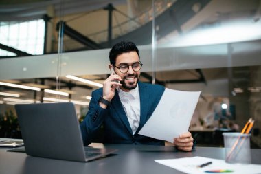 Takım elbiseli bir adam telefonda konuşurken gülümser ve çağdaş bir ofiste evraklara bakar. Çalışma alanı bir dizüstü bilgisayar ve düzenli kırtasiye malzemesi içeriyor.