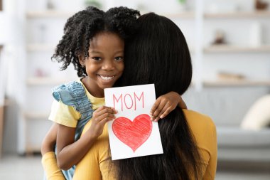 Annesini kucaklayan genç bir kız, elinde renkli bir kartla sevinçle ışıldıyor. 