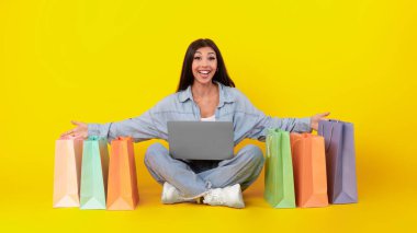 Genç bir kadın dizüstü bilgisayarını tutarken elinde renkli alışveriş torbalarıyla yerde bağdaş kurarak oturuyor. Mutlu ve meşgul görünüyor, canlı sarı bir arka plana karşı canlı bir sahne yaratıyor..