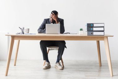 Bin yıllık siyahi bir ofis çalışanı elinde dizüstü bilgisayarla tahta bir masada oturur ve fazla mesaiden dolayı yorgunluk ve baş ağrısı belirtileri gösterir. Evrak işleri ve masasında stresli görünen bir bitki var..