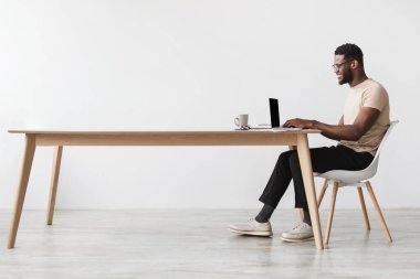 Genç siyah bir adam modern bir masada oturuyor, dizüstü bilgisayarına odaklanmış. Günlük kıyafetler giyer, parlak bir ofiste uzaktan çalışmaktan zevk alır. Kahve fincanı yakınlarda rahat bir atmosfer yaratıyor..