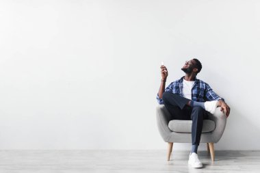 Neşeli genç siyah adam modern bir koltukta oturur, klimayı açmak için uzaktan kumanda kullanır. Basit beyaz bir duvara yaslanıp sakin bir atmosfer yaratıyor..