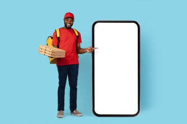 Mobile App. Cheerful Black Delivery Guy Holding Pizza Boxes And Pointing At Blank Phone With White Screen, African American Male Courier Posing Over Blue Studio Background, Mockup Image, Collage