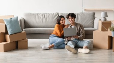 Sevgi dolu çift, yeni evleri için ev eşyaları almak için dizüstü bilgisayar kullanarak kutuları taşıyarak yerde oturuyor. Heyecanla birlikte geleceklerini planlıyorlar..