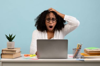 Siyahi bir kadın önünde dizüstü bilgisayarla masada oturuyor, şaşkın ve şaşkın görünüyor. Stres içinde başını tutuyor, okul projesi için mavi bir arkaplana karşı son teslim tarihi ile karşı karşıya..