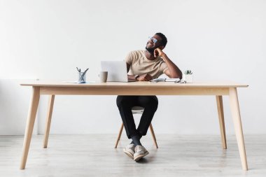 Neşeli bir siyah adam masasında oturuyor, online işine ara verirken düşüncelere dalmış. Parlak bir stüdyoda, beyaz bir arka planı olan anın tadını çıkarıyor..