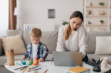 Bir anne dizüstü bilgisayarına odaklanırken, küçük çocuğu evde renkli yapı taşları ve sanat malzemeleriyle oynuyor. Sıcak oturma odası yumuşak ışıkla dolu..