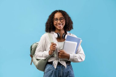 Gülümseyen siyah bir kız öğrenci elinde notlar ve dijital tablet tutarken kulaklık takıyor ve sırt çantası taşıyor..