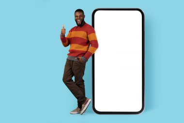 Çevrimiçi tanıtım. Neşeli siyahi adam Big Blank Smartphone 'un yanında duruyor ve başparmağını kaldırıyor, neşeli Afrikalı Amerikalı erkek cep telefonuna yaslanıyor Mockup İçin Beyaz Ekran, Tam Uzunluk