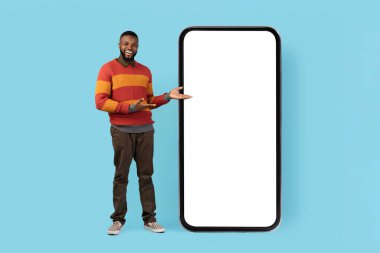 Yeni uygulama. Günlük kıyafetli neşeli siyah adam büyük boş telefonu işaret ediyor, gülümseyen Afro-Amerikan erkek cep telefonu beyaz ekranlı cep telefonu mobil web sitesi tasarımı, mavi arka plan, model.