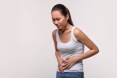 Beyaz bluzlu hasta bir Çinli kadın ishalden dolayı karnına dokunuyor. Gri bir stüdyo arkaplanından dolayı sıkıntılı görünüyor, fotokopi alanı mevcut..