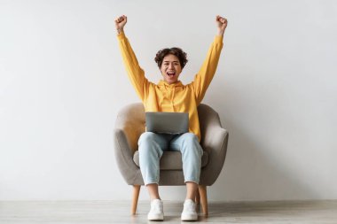 Neşeli genç Asyalı bir adam rahat bir koltukta oturur, heyecanla ellerini kaldırır ve bir anlık başarının tadını çıkarır. Dizüstü bilgisayarıyla çalışıyor. Parlak, beyaz bir stüdyoda..