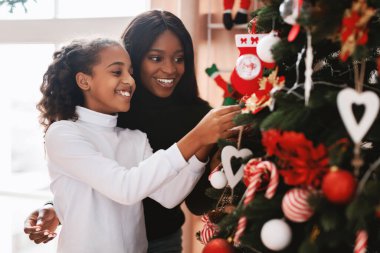 Neşeli bir siyahi kadın ve ergen bir kız sıcak oturma odalarında birlikte bir Noel ağacı süslüyorlar. Renkli süsler asarlar, kahkahaları ve bayram neşesini paylaşırlar..