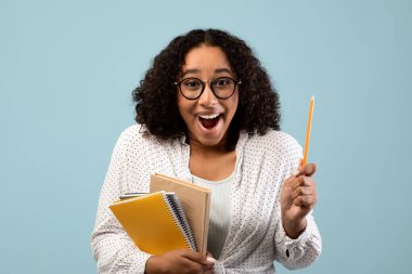 Elinde çalışma materyalleri tutan heyecanlı siyah kız öğrenci bir ilham anı yaşıyor. Yaratıcı düşüncelerini mavi bir stüdyo ortamında dışa vurarak geniş bir şekilde gülümsüyor..