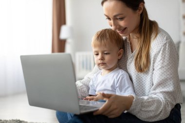 Bir anne parlak bir oturma odasının zemininde oturur ve küçük çocuğuna dizüstü bilgisayar kullanmasında rehberlik eder. Güneş ışığı pencereden akar ve sıcak bir atmosfer yaratır..