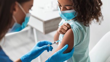 Genç siyahi bir kız klinikte otururken bir sağlık çalışanı Covid-19 aşısını yönetiyor. Her ikisi de maske takıyor, bağışıklık kazanırken güvenliği sağlıyorlar..