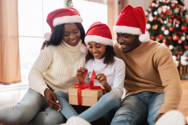 Neşeli siyah bir kız, hediye paketini açarken, Noel Baba şapkası takan mutlu anne ve babasıyla yerde oturuyor. Süslü Noel ağacı oturma odasındaki şenlikli atmosfere katkı sağlar..