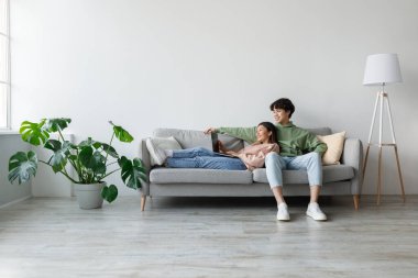 Neşeli genç Asyalı çift oturma odalarındaki bir kanepede rahatlıyor ve bir video mesajı aracılığıyla başkalarıyla bağlantı kurmak için dizüstü bilgisayar kullanıyorlar. Atmosfer rahat ve doğal ışıkla davetkar..