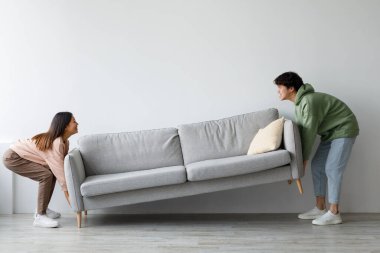 Genç Asyalı çift oturma odalarında gri bir kanepeyi kaldırırken gülümsüyor. Dairelerindeki mobilyaları değiştirmek için birlikte çalışıyorlar, sürecin tadını çıkarıyorlar..