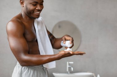Neşeli bir Afrikalı Amerikalı adam modern bir banyoda üstsüz duruyor, traş rutinine ve kişisel bakımına hazırlanırken eline tıraş köpüğü sürüyor..