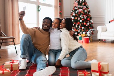 Üç çocuklu neşeli siyah aile, Noel 'i birlikte oturma odasında kutluyor. Etrafı şenlikli süslemeler, hediyeler ve Noel ağacı ile çevriliyken selfie çekiyorlar..