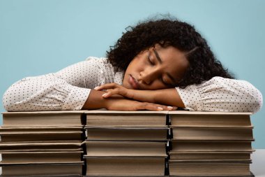 Yorgun bir Afrikalı Amerikalı kız öğrenci kafasını kitap yığınlarının üzerinde dinlendiriyor. Yoğun sınav hazırlığından bitkin düştüğü aşikâr. Mavi stüdyo arka planı yorgunluk ve odaklanma duygusunu arttırır..