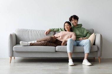 Neşeli bir Asyalı çift rahat bir koltukta oturup kameraya gülümsüyor. Rahatlar, boş vakitlerinde rahat oturma odalarında birbirlerine eşlik ederler..