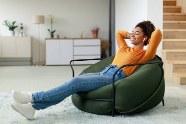 Genç bir kadın evdeki modern bir sandalyeye oturmuş kulaklık takıyor ve gülümsüyor. Turuncu bir kazak ve kot pantolon giyiyor ve bir anlık huzurun tadını çıkarıyor..