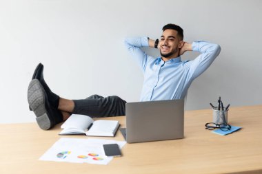Gülümseyen genç bir Arap, masadaki sandalyesine yaslanır ve bacaklarını masaya yaslar. Dizüstü bilgisayar kullanırken mutlu ve rahat görünüyor ve işteki molasının tadını çıkarıyor..