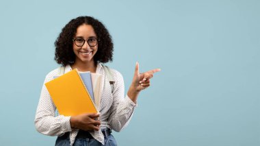 Sırt çantalı neşeli siyah bir kız öğrenci çalışma materyallerini elinde tutuyor ve reklamınız için boş bir alanı işaret ediyor. Açık mavi arka planda eğitim tekliflerini paylaşmaya hazır..