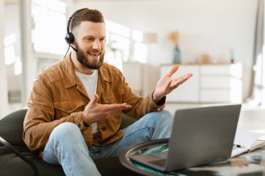 Kulaklık takan bir adam rahat bir oturma odasında oturup dizüstü bilgisayarındaki biriyle canlı bir şekilde tartışır. Doğal ışık odayı dolduruyor..