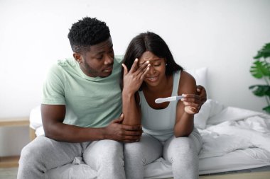 Bir çift, hamilelik testine bakarken karışık duygular göstererek bir yatakta yan yana oturur. Oda hafifçe aydınlanıyor, beklentiyle dolu samimi bir atmosfer yaratıyor..