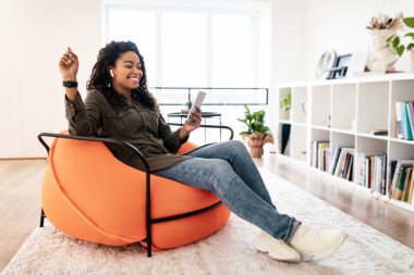 Genç bir kadın modern bir oturma odasında parlak turuncu bir fasulye çuvalında rahat bir şekilde oturuyor, akıllı telefonuna bakarken gülümsüyor. Sıradan giyinmiş, evde vakit geçiriyor..