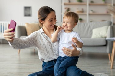 Bir anne ve küçük oğlunun selfie çekerken gülümsediği neşeli bir an. Oturma odasındaki rahat atmosfer bağlantılarını güçlendiriyor..