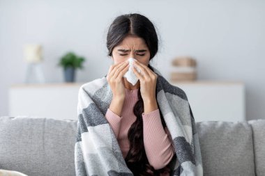 Ekose giyinmiş üzgün bir Hintli kadın oturma odasında kanepede oturuyor, grip ve ateş yüzünden acı çekiyor. Battaniyeye sarılı bir şekilde burnunu peçeteye sümkürerek rahatsızlığını gösteriyor..