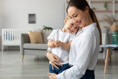 Bir anne, genç oğlunu parlak bir oturma odasında tutarken sıcak bir şekilde gülümser. Çocuk sevgiyle ona yaslanıyor, mutluluk saçıyor. Yumuşak mobilyalar rahat atmosferi güçlendirir..