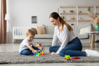 Genç bir çocuk yumuşak bir halıya oturur, annesi yanında diz çökerken renkli bloklar dizer, onunla meşgul olur. Konforlu oturma odası doğal ışık ve modern dekorlarla dolu..