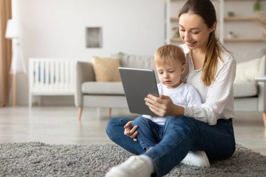 Bir anne ve küçük oğlu yumuşak bir halıya oturup tablete bakıyorlar. Sıcak güneş ışığı, rahat oturma odasını dolduruyor ve rahat bir atmosfer yaratıyor..