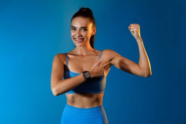 Bir jimnastikçi kadın mavi arka plandaki kaslarını gösterirken gülümsüyor. Sıkı çalışmasını ve kendini spora adadığını gösteriyor. Diğerlerini spor amaçlarına ulaşmaları için motive ediyor..