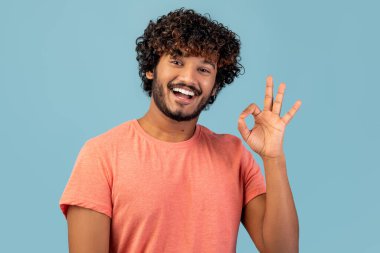 Pembe tişörtlü mutlu ve yakışıklı bir genç doğu erkeği gülümser ve açık mavi arka plana karşı iyi bir jest yapar. Kendinden emin görünüyor ve olumlu bir şey öneriyor..