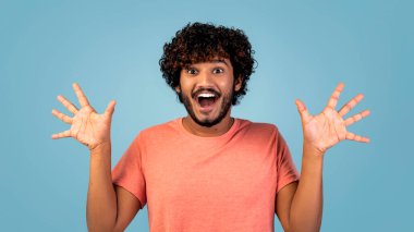 Kıvırcık saçlı Hindu bir adam mavi bir stüdyoda duruyor, ellerini kaldırıyor ve heyecan gösteriyor. Yüzündeki ifade neşe ve şaşkınlık veriyor, neşeli bir anı yakalıyor..