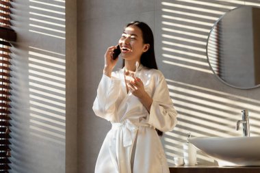 İpek bornozlu neşeli bir bayan telefonda konuşurken dişlerini fırçalıyor. Aynaya gülümsüyor, yumuşak ışıkla dolu şık bir banyoda sabah rutininin tadını çıkarıyor..