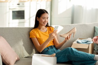 Heyecanlı bir kadın kanepesinde otururken içinde yeni ayakkabılar olan bir paket açıyor. Çevrimiçi mağazayla ilgili olumlu görüşleri paylaşmak için akıllı telefonuyla fotoğraf çekiyor..