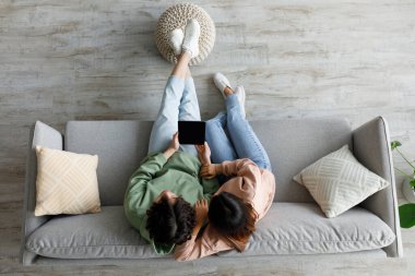 Asyalı çift rahat bir koltukta oturuyor, ellerinde boş ekranlı bir tablet var. Rahatlar ve meşguldürler, bir ev ortamında tasarım veya uygulama promosyonları için fırsat sağlarlar..