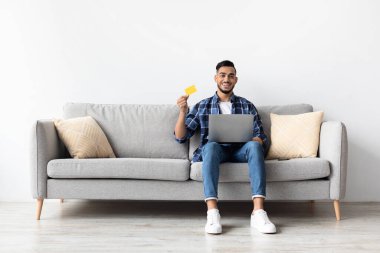 Kanepede rahatça oturan gülümseyen bir adam elinde kredi kartı tutarken dizüstü bilgisayar kullanıyor. Çevrimiçi alışveriş ve para transferi ile meşgul, rahat bir ev ortamı sergiliyor.