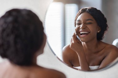 Üstsüz, milenyum siyahi bir kadın banyo aynasının önünde pamuk ped ve güzellik ürünü kullanarak makyajını zevkle temizliyor. O, rutini sırasında yansımasına gülümsüyor..