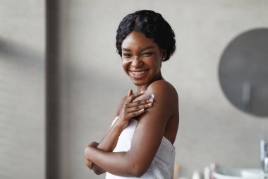 Neşeli genç siyah bir bayan dinlendirici bir duştan sonra omzuna vücut losyonu sürerken kameraya gülümsüyor. Temiz, parlak bir banyoda yumuşak bir havluya sarılmış..