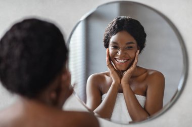 Parlak bir banyoda, neşeli genç siyah bir kadın yüzüne su veren bir ürün sürerken gülümsüyor..