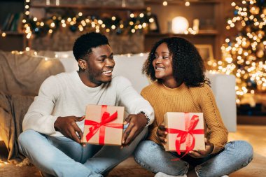 Genç siyahi çift, bayram süslemeleri ve ışıklarıyla dolu sıcak, şenlikli bir oturma odasında dizlerinin üstüne çöküp gülümseyerek Noel hediyelerini değiş tokuş ediyorlar..