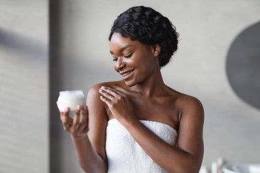 Genç siyah bir kadın modern bir banyoda sabah rutininin tadını çıkarıyor. Omzuna vücut losyonu sürerken gülümsüyor, duştan yeni çıkmış ve havluya sarılmış..